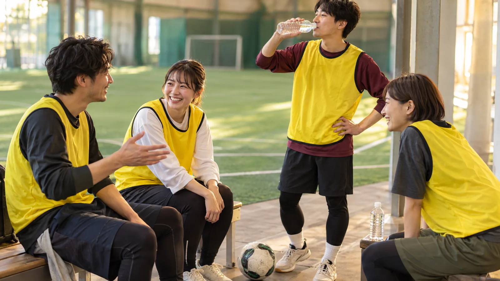 屋内フットサルコート脇のベンチで、黄色のビブスを着た4人が試合の合間に笑顔で話し、1人が水を飲んでいる場面